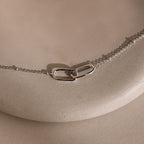 Linked Pendant Necklace in Silver showcases two interlocking oval links on a sleek silver chain, elegantly displayed on a smooth beige surface.