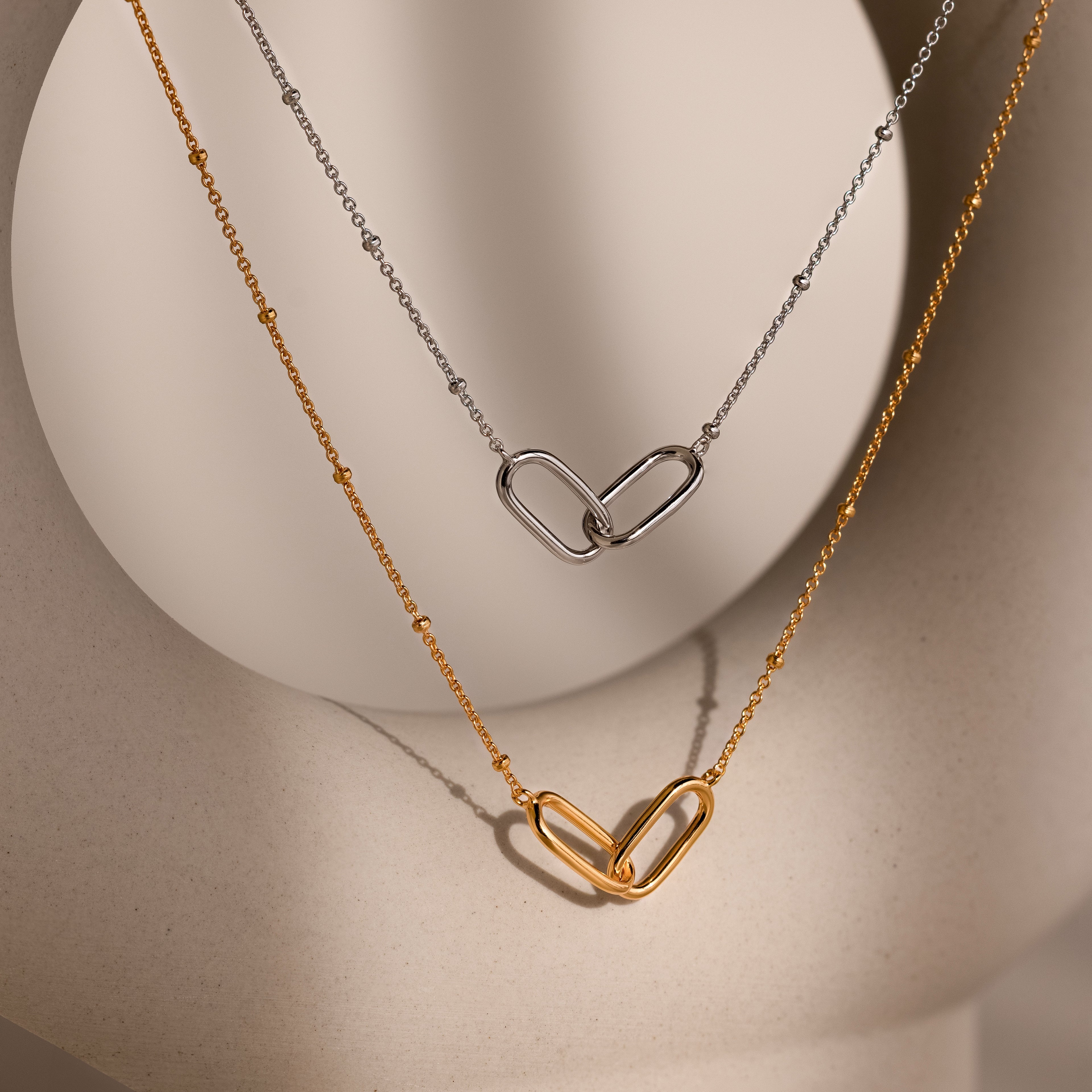 The Linked Pendant Necklace in Silver with a delicate gold chain is displayed on a neutral, curved surface.