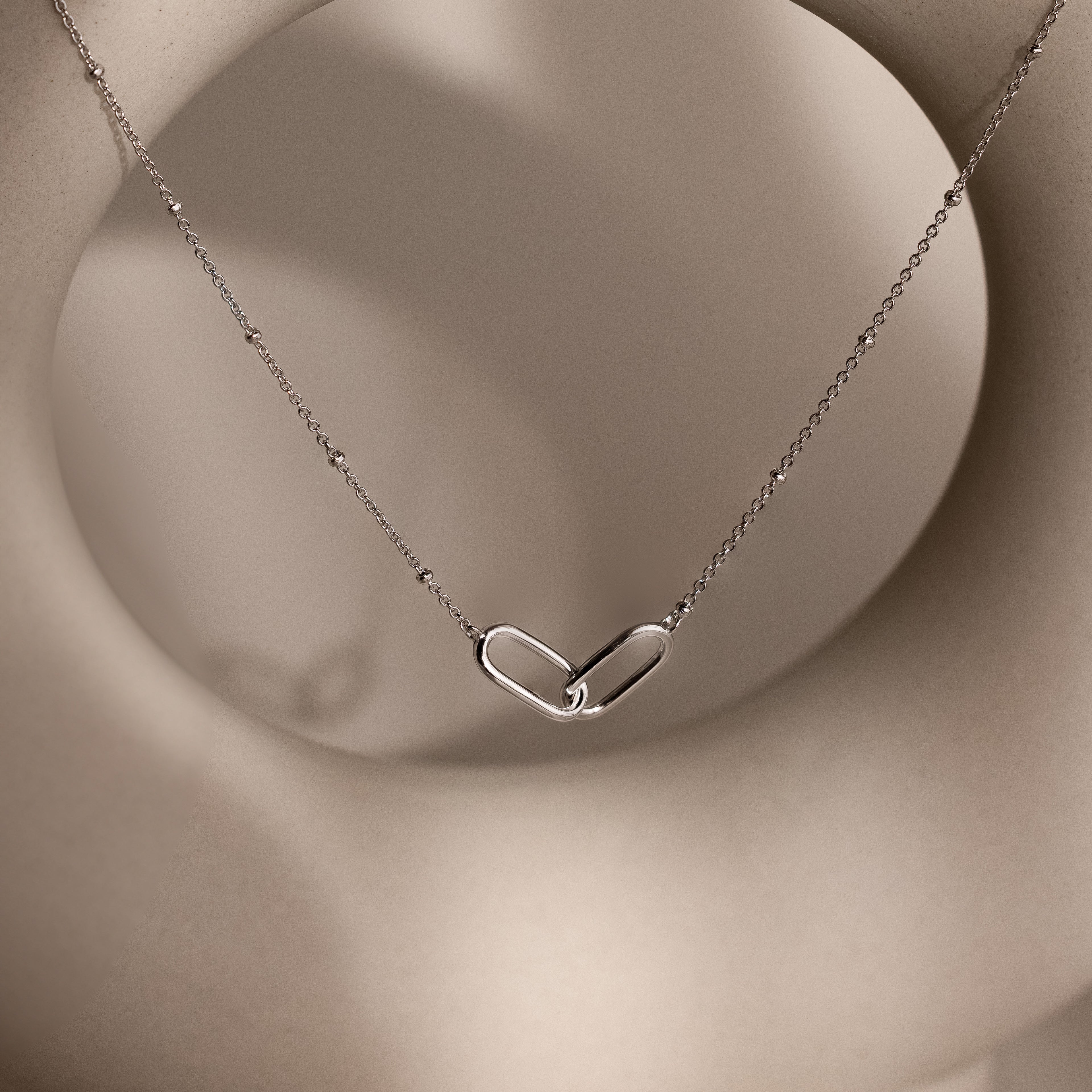 Linked Pendant Necklace featuring a heart-shaped linked pendant on a delicate chain, shown against a soft beige circular background.