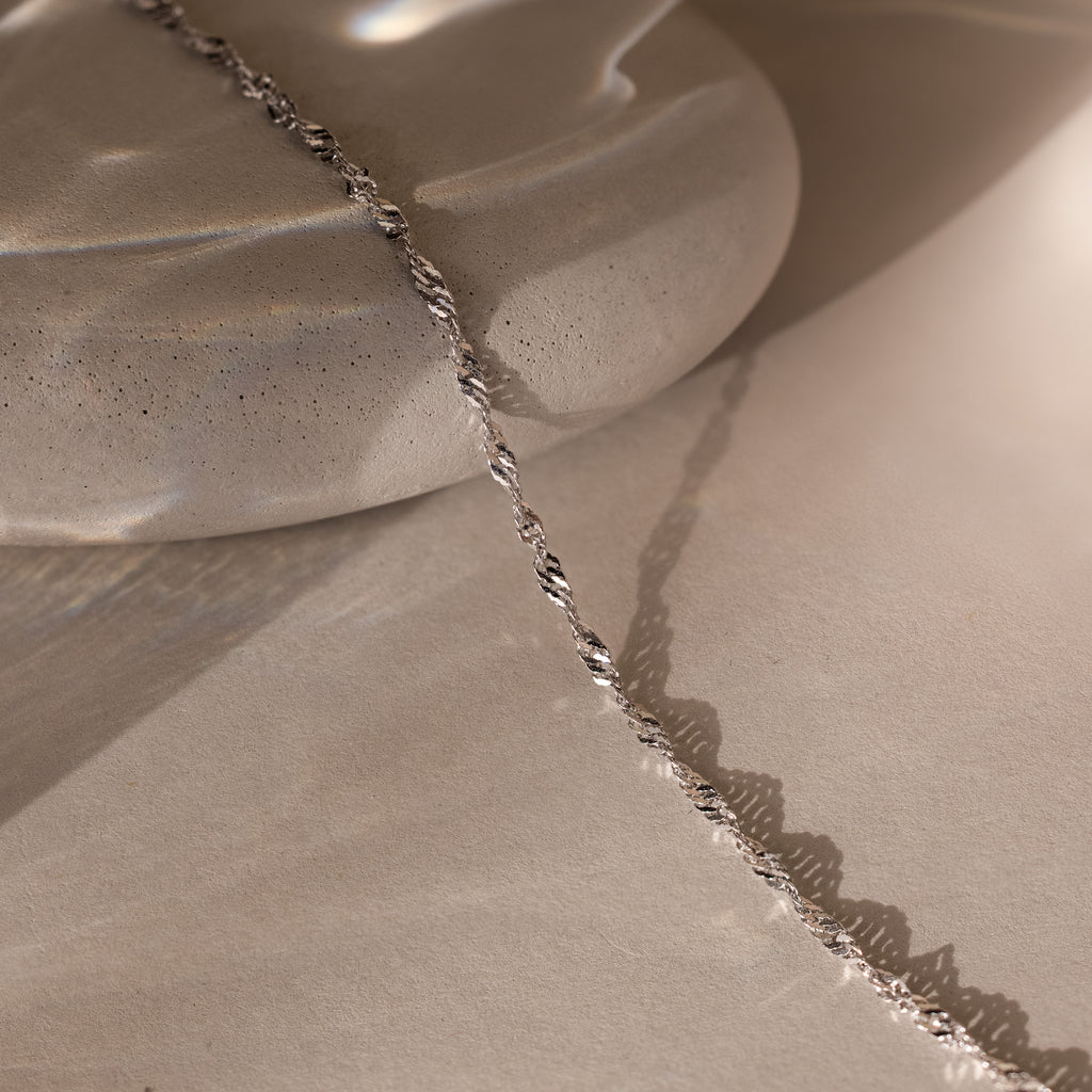 The Singapore Twist Necklace in Silver is displayed elegantly on a smooth, light stone surface.