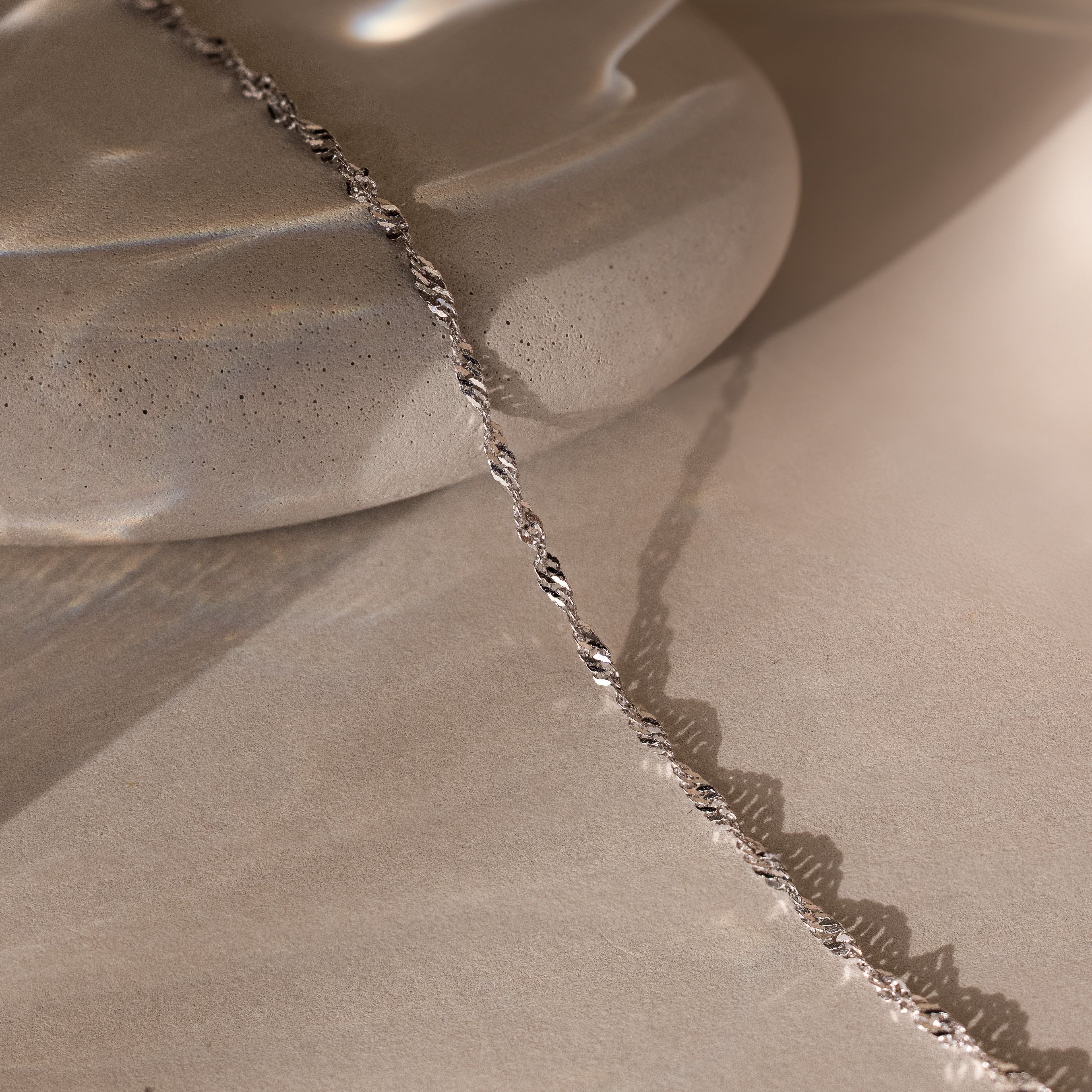 The Singapore Twist Necklace in Silver is displayed elegantly on a smooth, light stone surface.