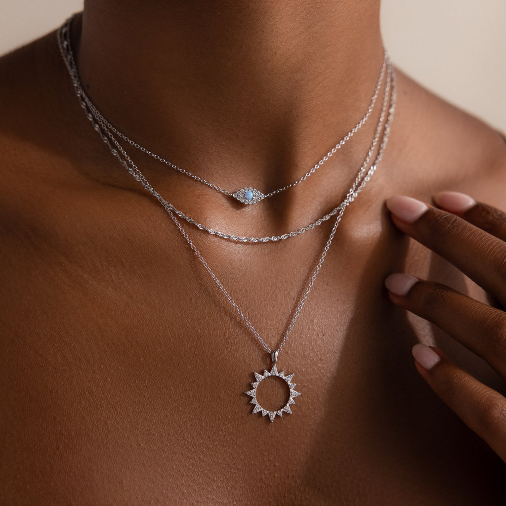 A close-up of a person wearing layered silver necklaces, including a sun pendant amulet on a rope chain and the Singapore Twist Necklace in Silver with a small round gem.