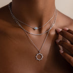 A close-up of a person wearing layered silver necklaces, including a sun pendant amulet on a rope chain and the Singapore Twist Necklace in Silver with a small round gem.