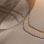 The Singapore Twist Necklace is displayed on a smooth stone surface in gold and silver with soft lighting and gentle shadows.