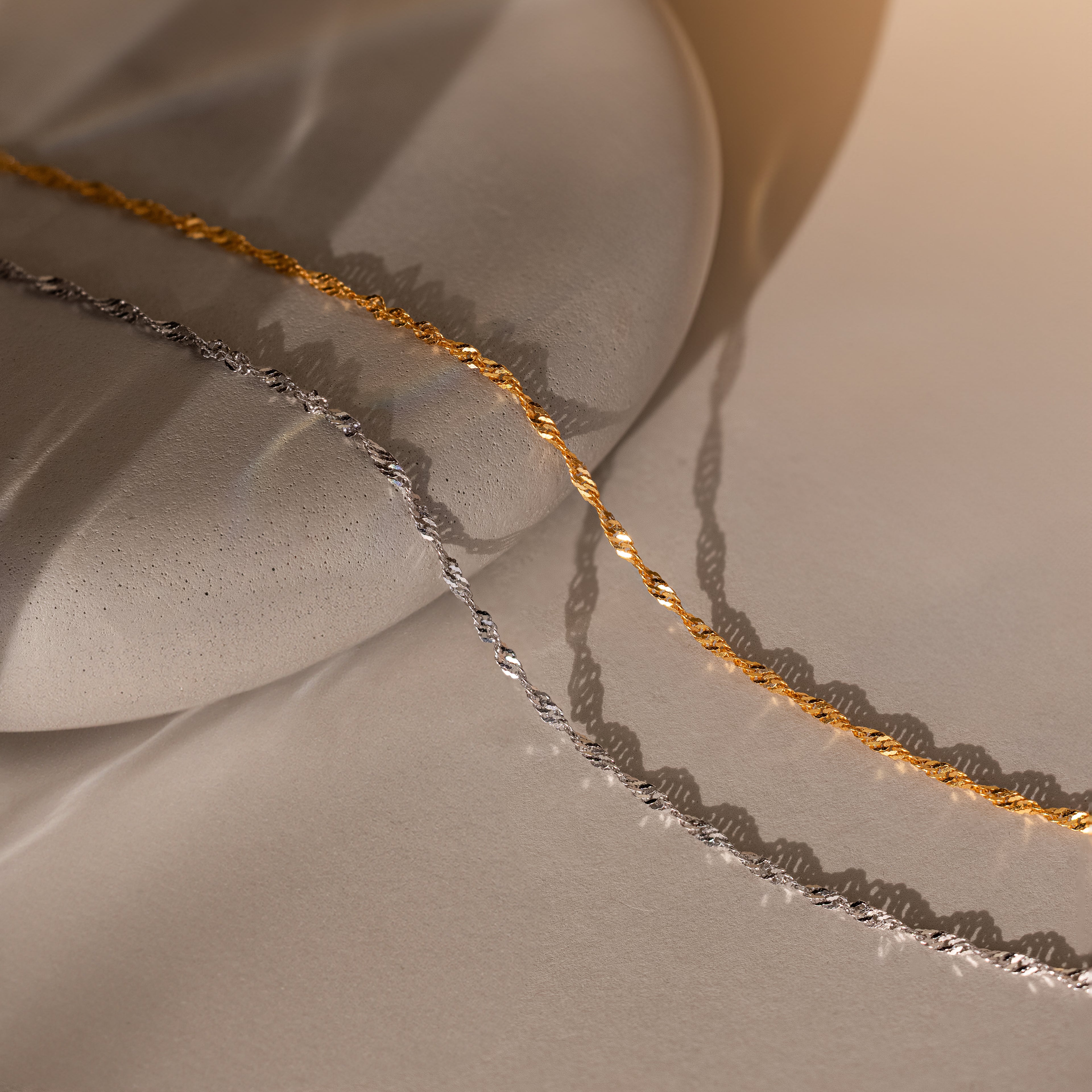 The Singapore Twist Necklace is displayed on a smooth stone surface in gold and silver with soft lighting and gentle shadows.
