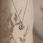 Bee Coin Necklace in Silver featuring a delicate bee design, ideal for layering. Displayed on a textured background with leafy shadows.