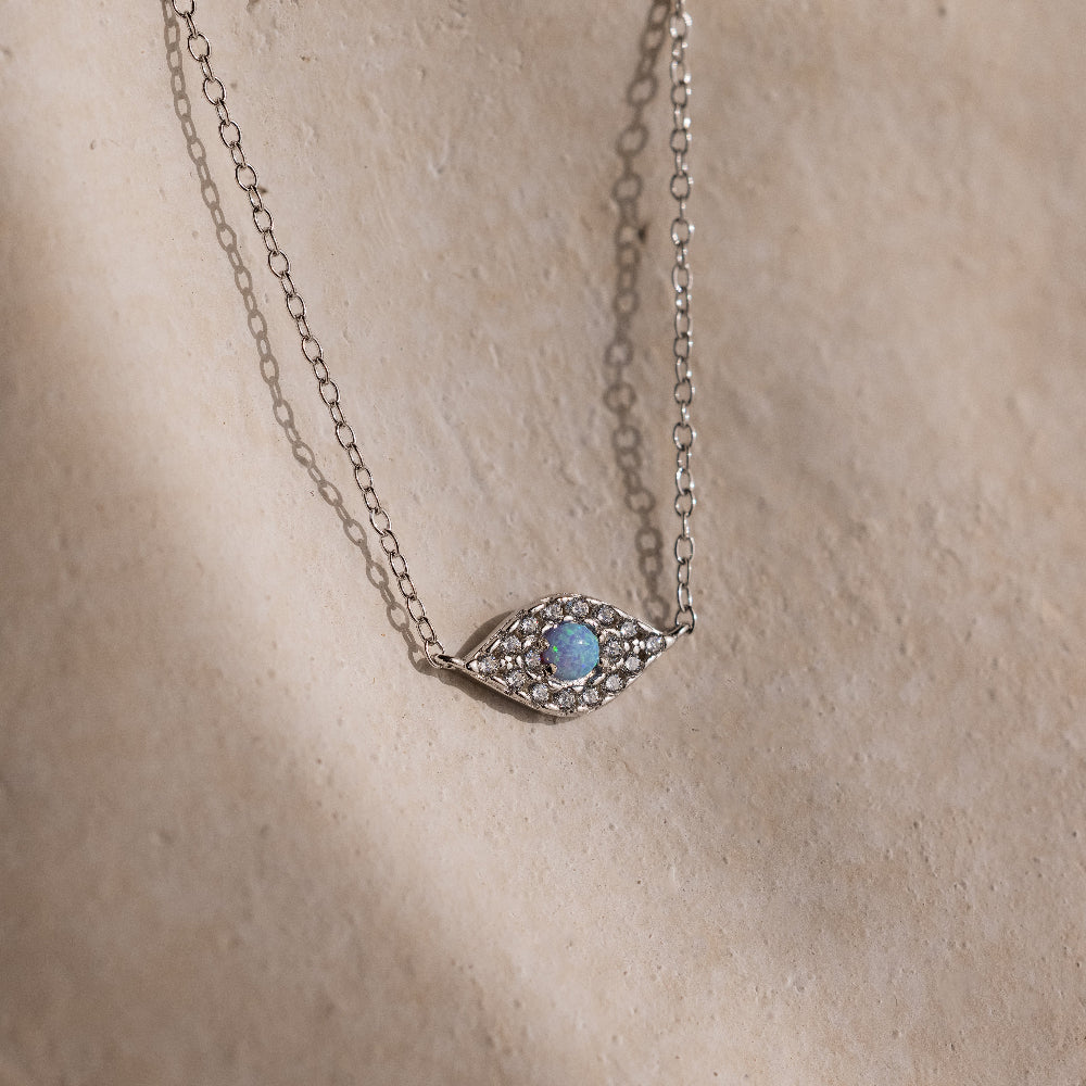 The Opal Eye Necklace in Silver features a blue stone and small clear crystals, creating a striking lucky talisman, showcased on a beige surface.