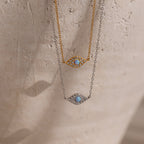 The Opal Eye Necklace in silver and gold features a blue stone center in an eye-shaped pendant—an elegant talisman, perfect for adding luck and style on a beige background.