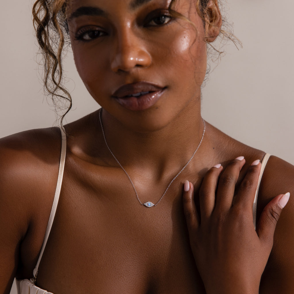 A woman touches her collarbone, wearing the Opal Eye Necklace—a delicate silver piece with a blue opal pendant—captured in soft lighting for a subtle, lucky talisman look.