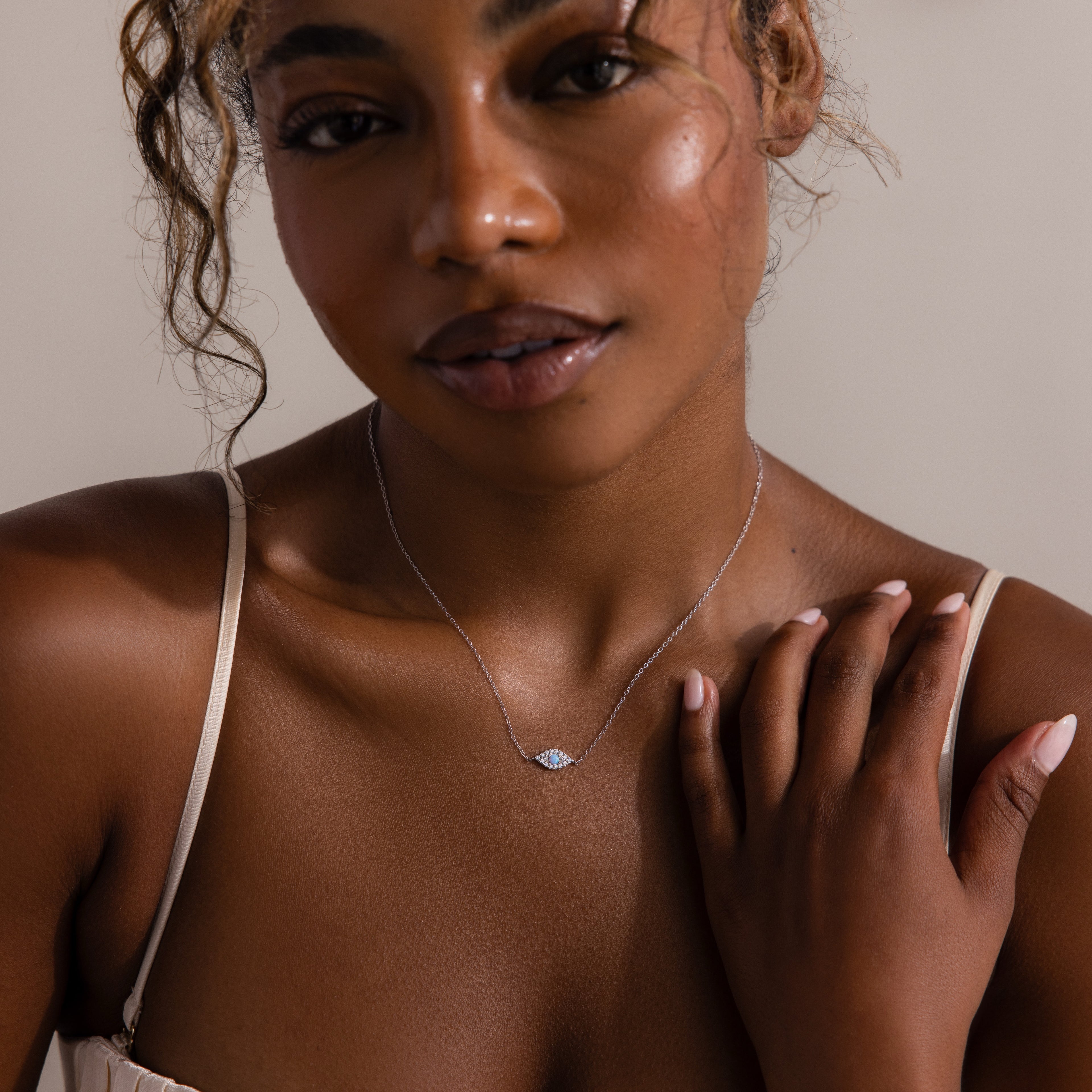 A woman touches her collarbone, wearing the Opal Eye Necklace—a delicate silver piece with a blue opal pendant—captured in soft lighting for a subtle, lucky talisman look.