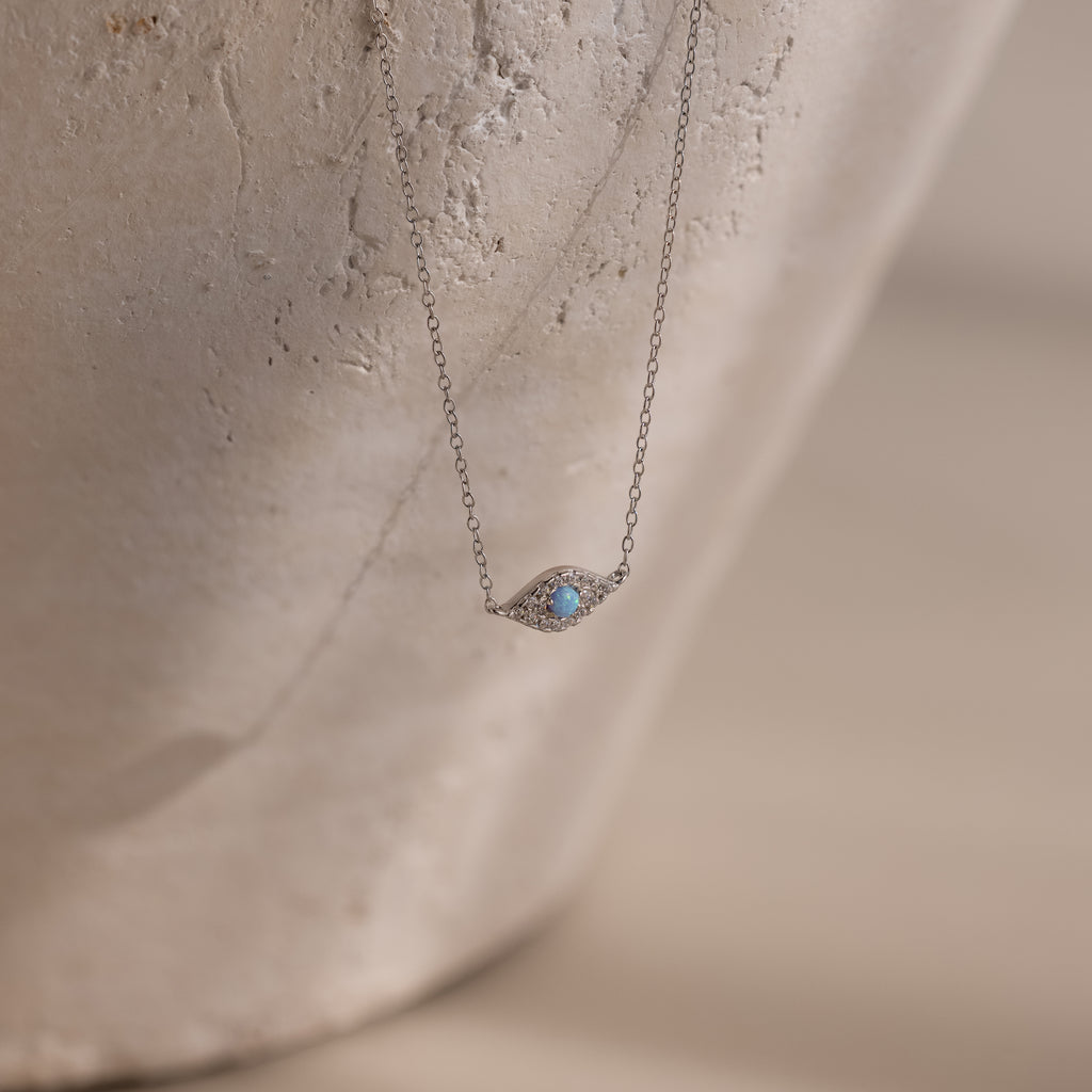 The Opal Eye Necklace in Silver features a delicate blue gemstone pendant, shown on a textured, light background—an elegant piece perfect as your lucky talisman.