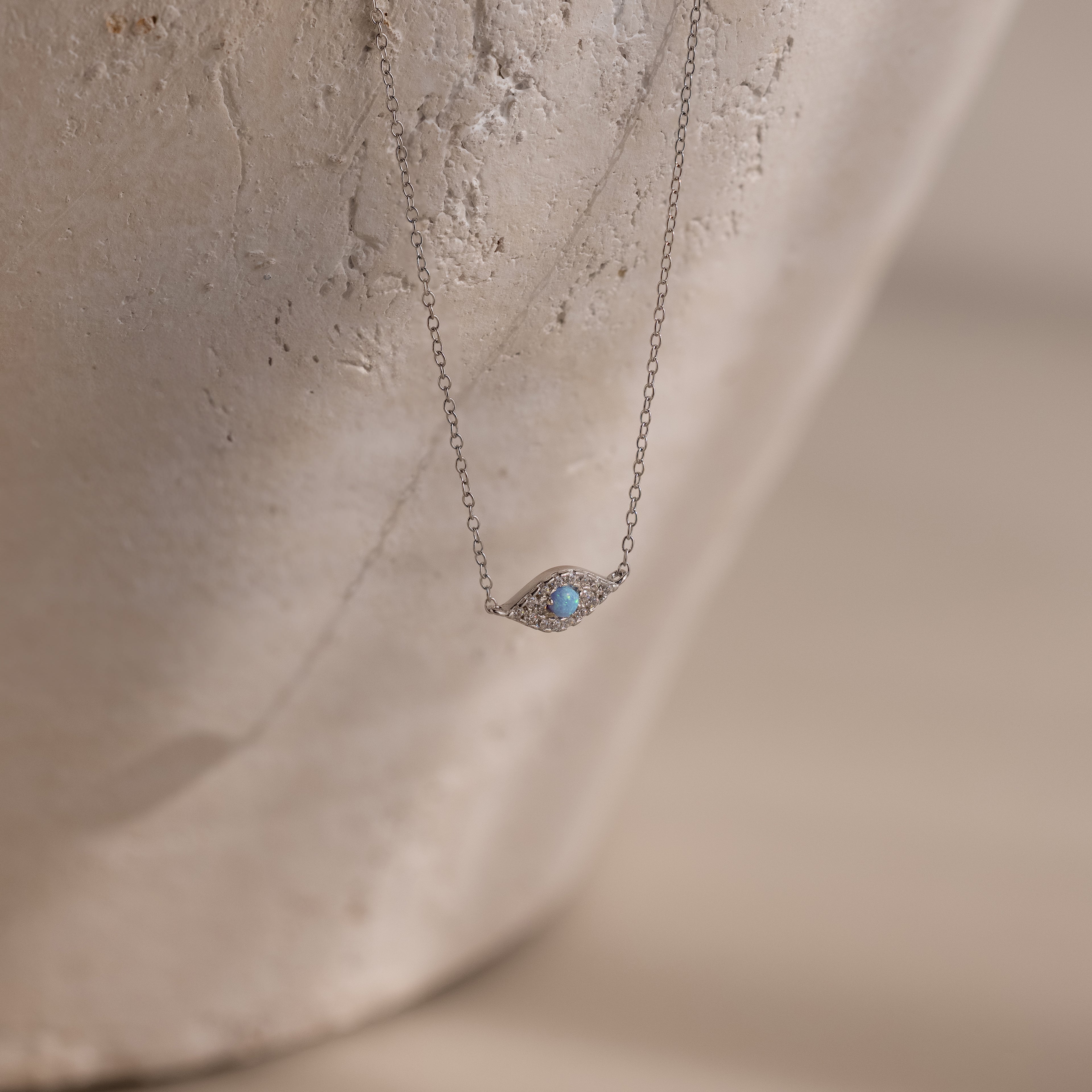 The Opal Eye Necklace in Silver features a delicate blue gemstone pendant, shown on a textured, light background—an elegant piece perfect as your lucky talisman.