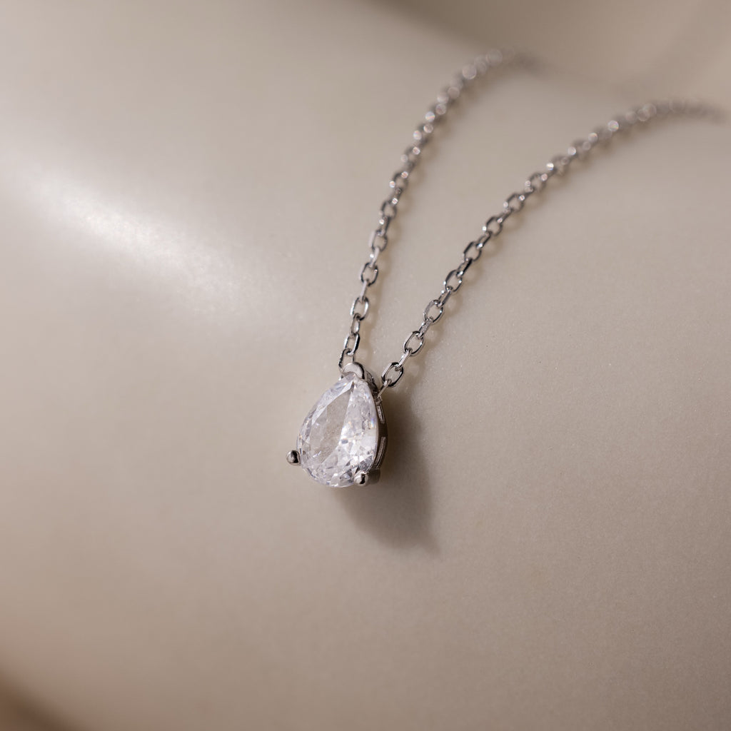The Rae Diamond Necklace in Silver, featuring a teardrop-shaped diamond pendant, rests on a smooth beige surface.