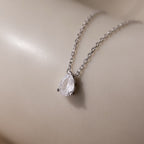 The Rae Diamond Necklace in Silver, featuring a teardrop-shaped diamond pendant, rests on a smooth beige surface.