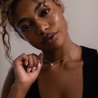 A woman with curly hair in a black top holds the Rae Diamond Necklace in Silver, featuring a teardrop-shaped diamond, as she looks softly at the camera.