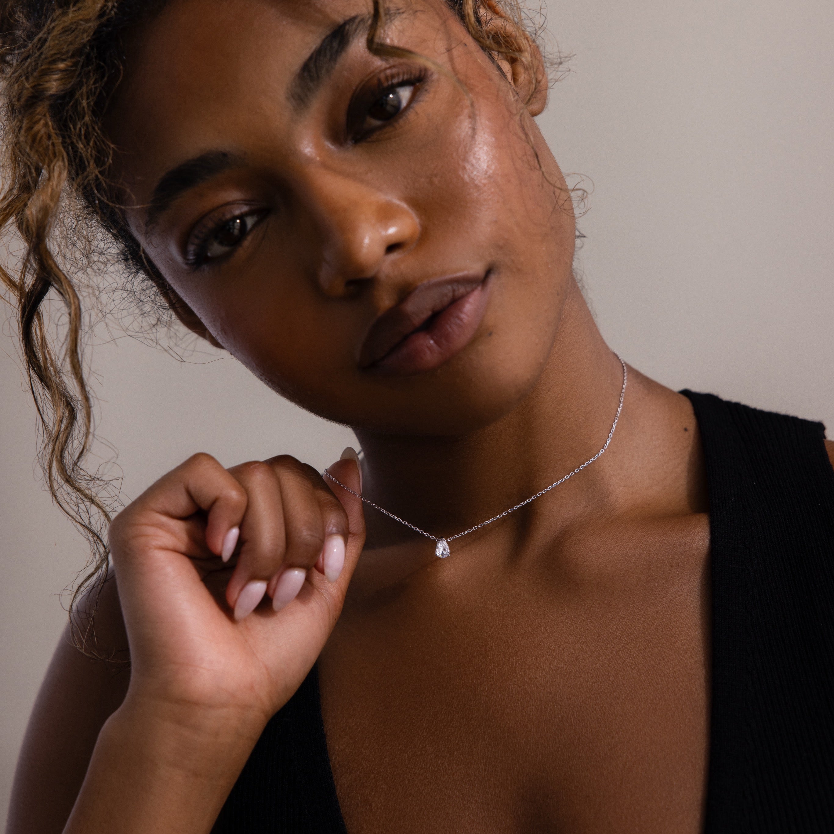 A woman with curly hair in a black top holds the Rae Diamond Necklace in Silver, featuring a teardrop-shaped diamond, as she looks softly at the camera.