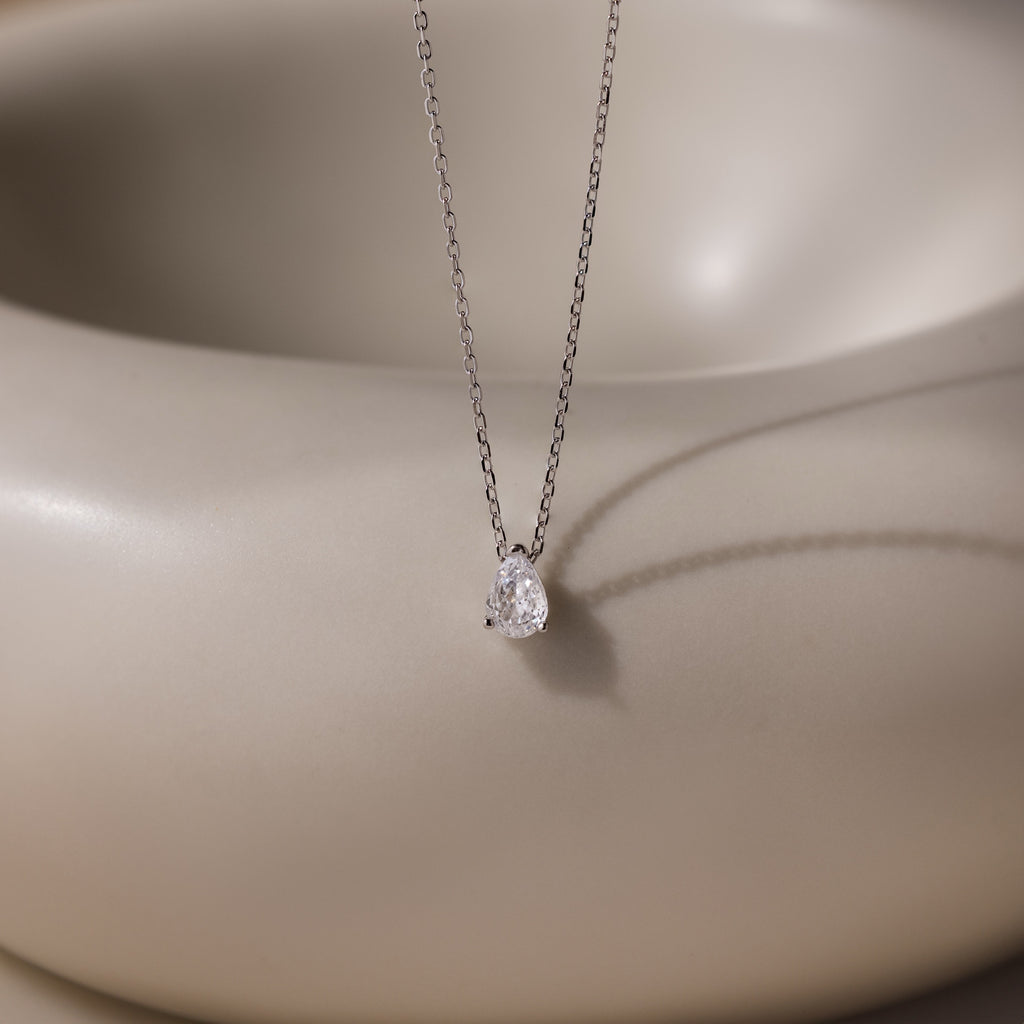 The Rae Diamond Necklace features a delicate silver chain with a teardrop diamond pendant, showcased on a smooth beige background—an elegant and exquisite jewelry piece.