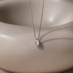 The Rae Diamond Necklace features a delicate silver chain with a teardrop diamond pendant, showcased on a smooth beige background—an elegant and exquisite jewelry piece.