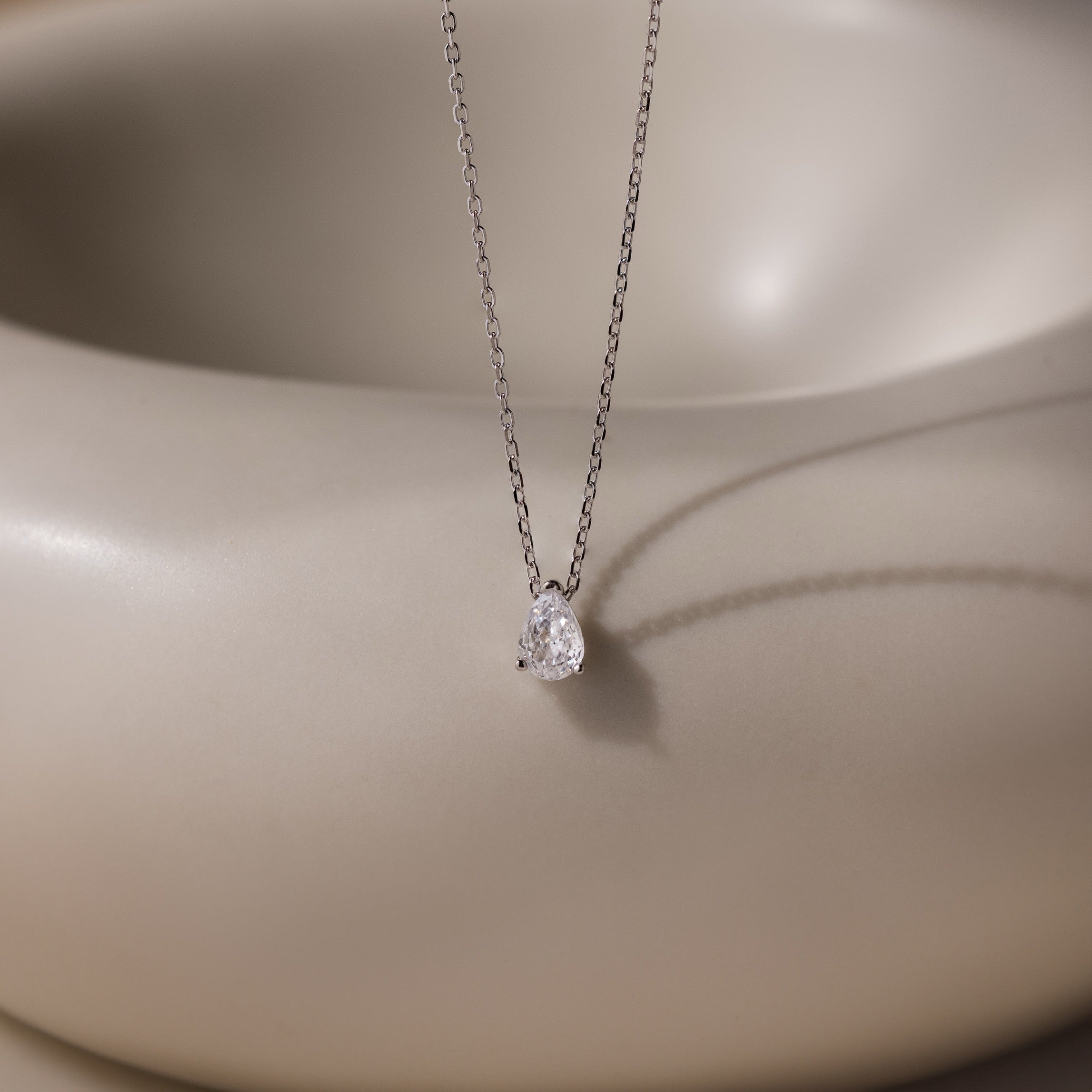 The Rae Diamond Necklace features a delicate silver chain with a teardrop diamond pendant, showcased on a smooth beige background—an elegant and exquisite jewelry piece.