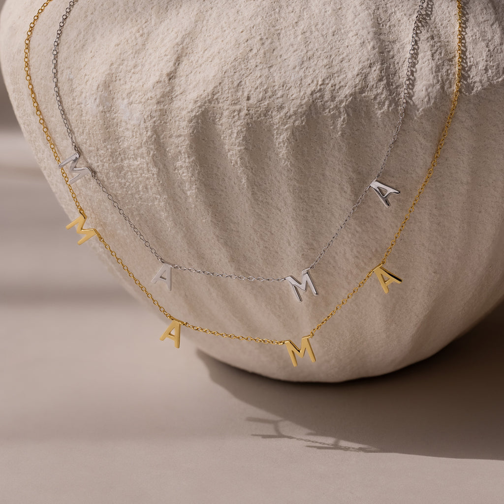 The Mama Letter Necklace in Silver and Gold, elegantly draped over a textured white vase, is a stylish tribute to motherhood and makes a thoughtful gift for any mom.