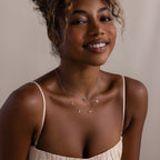 Woman in a cream pleated dress wears layered silver necklaces, including the Forever Pave Heart Necklace in Silver, against a neutral background.
