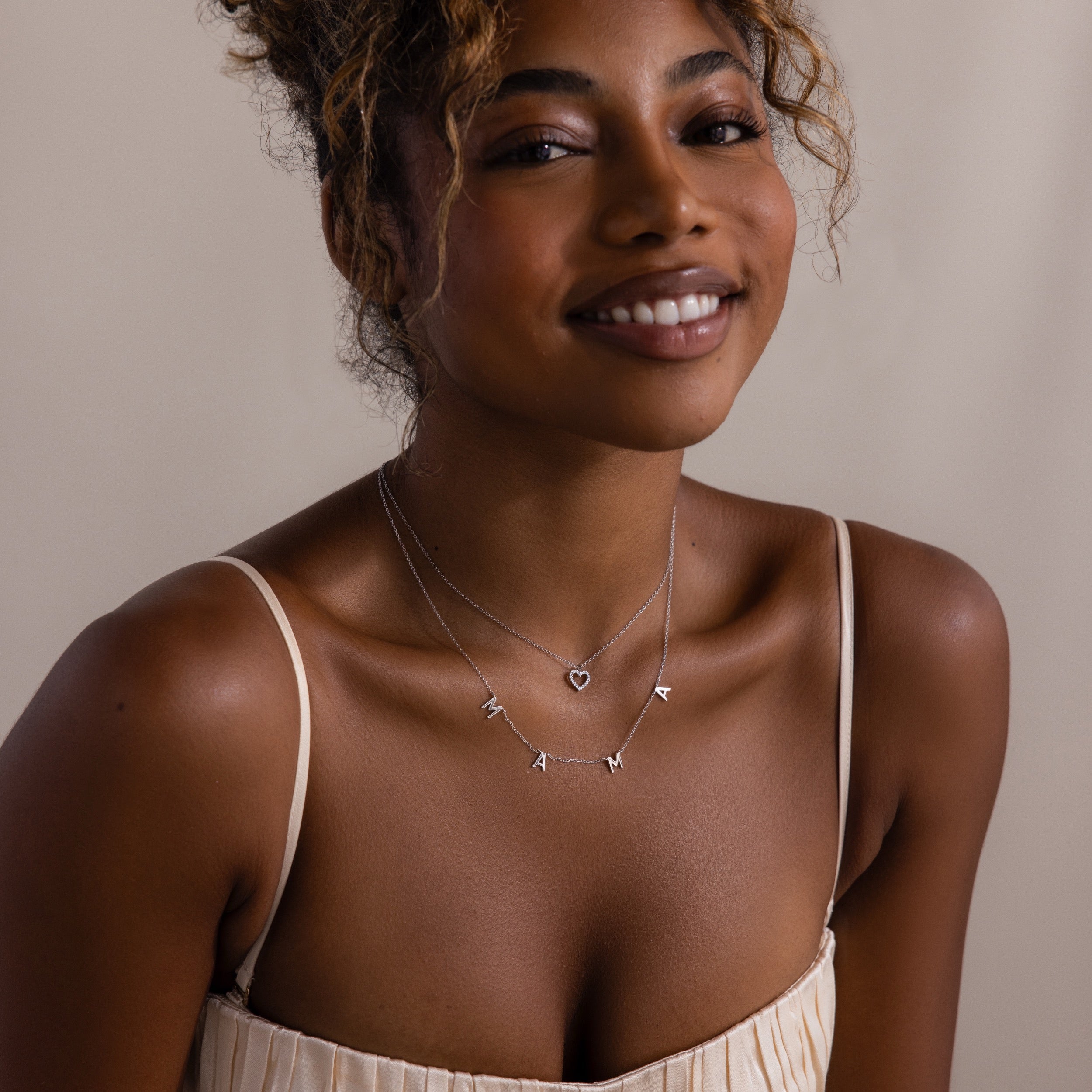 Woman in a cream pleated dress wears layered silver necklaces, including the Forever Pave Heart Necklace in Silver, against a neutral background.