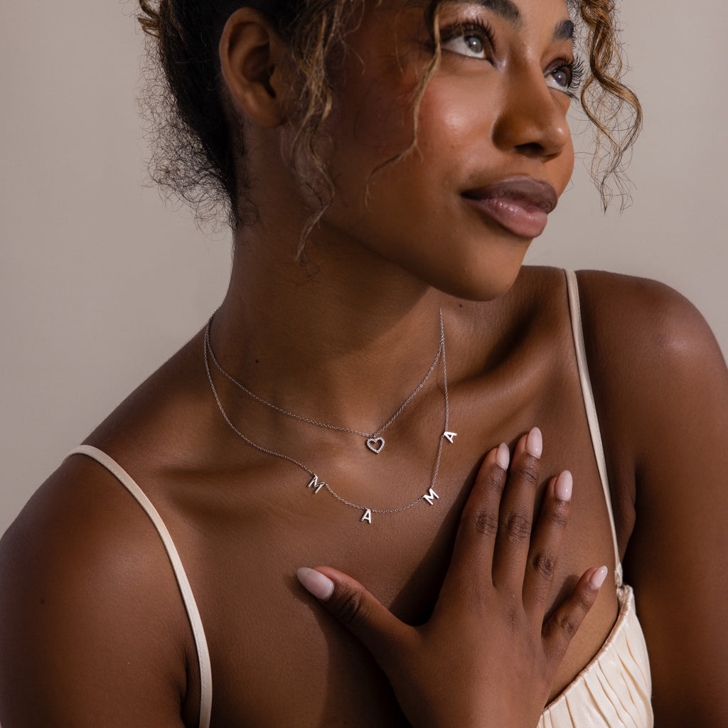 A woman wearing the Mama Letter Necklace touches her chest and smiles softly, celebrating motherhood with this meaningful gift.