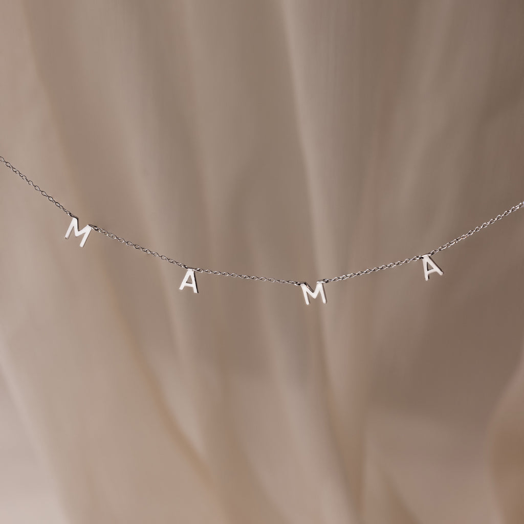The Mama Letter Necklace in Silver, featuring silver letter charms spelling MAMA, makes a perfect mother gift and is displayed against a soft beige background.
