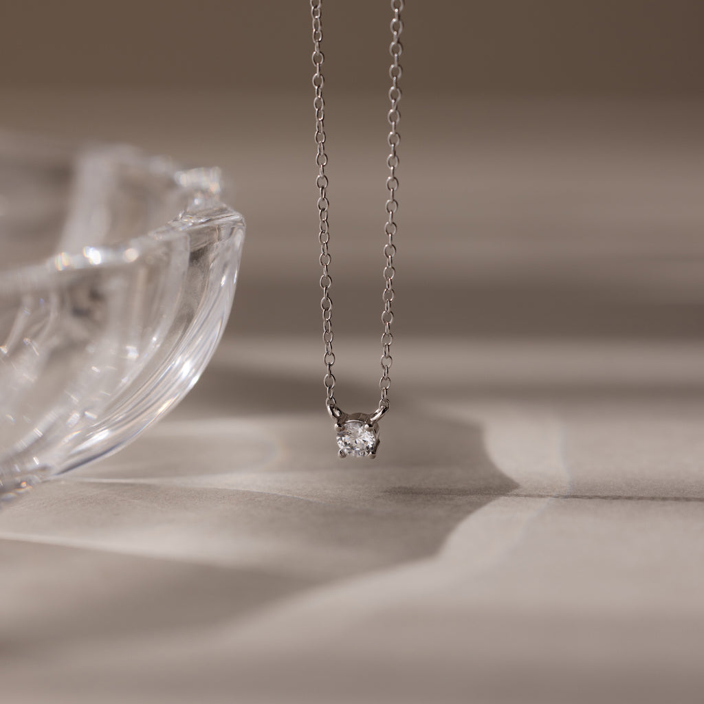 The Dainty Diamond Necklace in Silver, featuring a solitaire diamond pendant, is displayed beside a clear glass dish on a beige surface—an elegant Christmas gift choice.