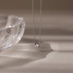 The Dainty Diamond Necklace in Silver, featuring a solitaire diamond pendant, is displayed beside a clear glass dish on a beige surface—an elegant Christmas gift choice.