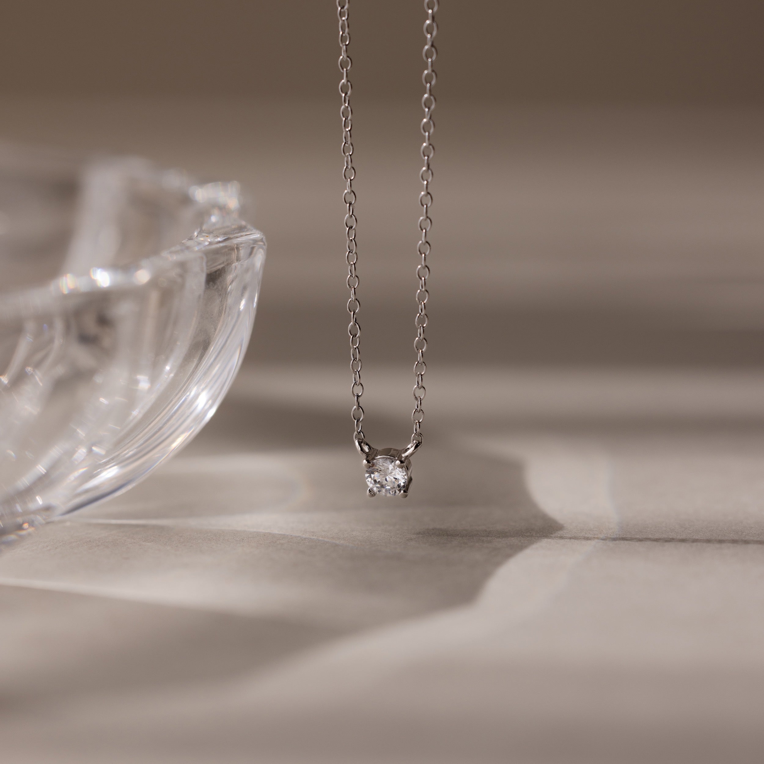 The Dainty Diamond Necklace in Silver, featuring a solitaire diamond pendant, is displayed beside a clear glass dish on a beige surface—an elegant Christmas gift choice.