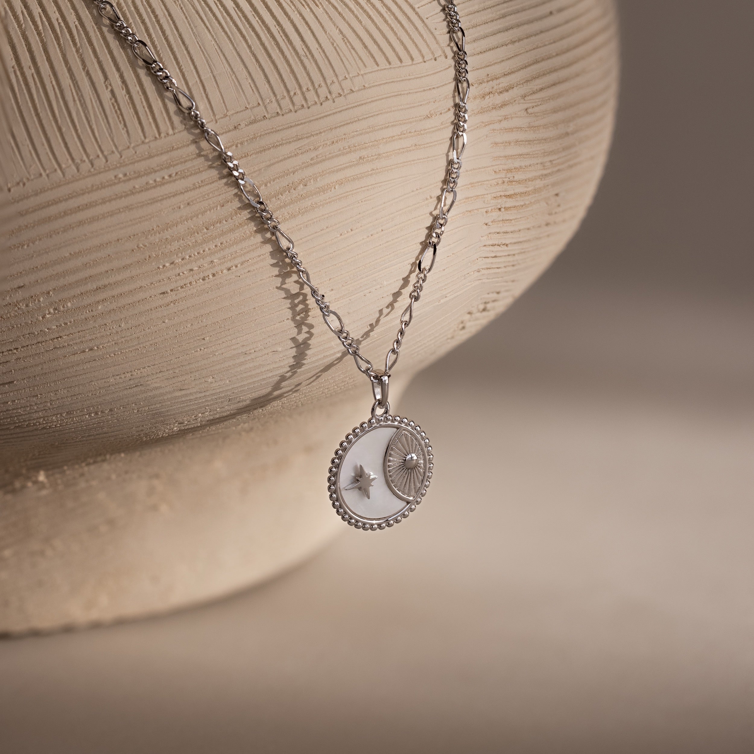 The Gabi Coin Necklace features a round sun and star pendant, displayed on a textured ceramic vase.