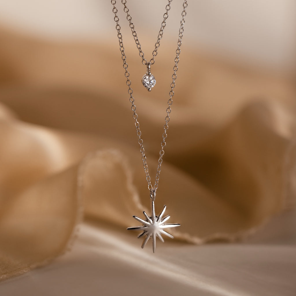 The Laura Starburst Duo Necklace in Silver, featuring a dazzling starburst pendant and diamond accent, is displayed on soft beige fabric.