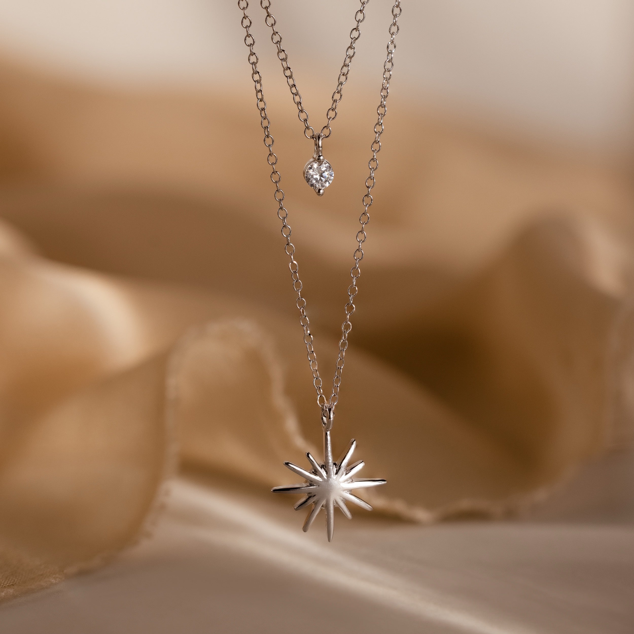 The Laura Starburst Duo Necklace in Silver, featuring a dazzling starburst pendant and diamond accent, is displayed on soft beige fabric.