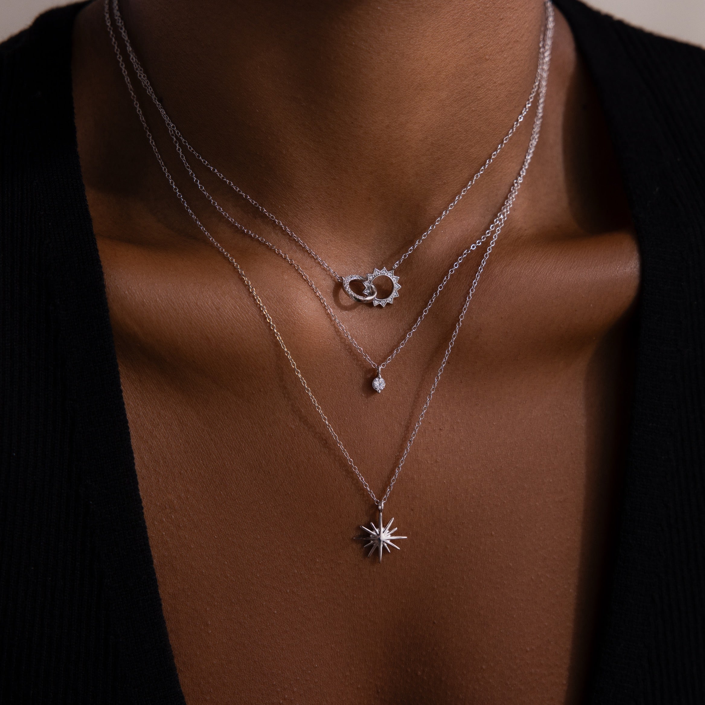 A person wears three layered silver necklaces, featuring the Sun  Moon Eternity Necklace, a celestial circle necklace, and a starburst pendant.
