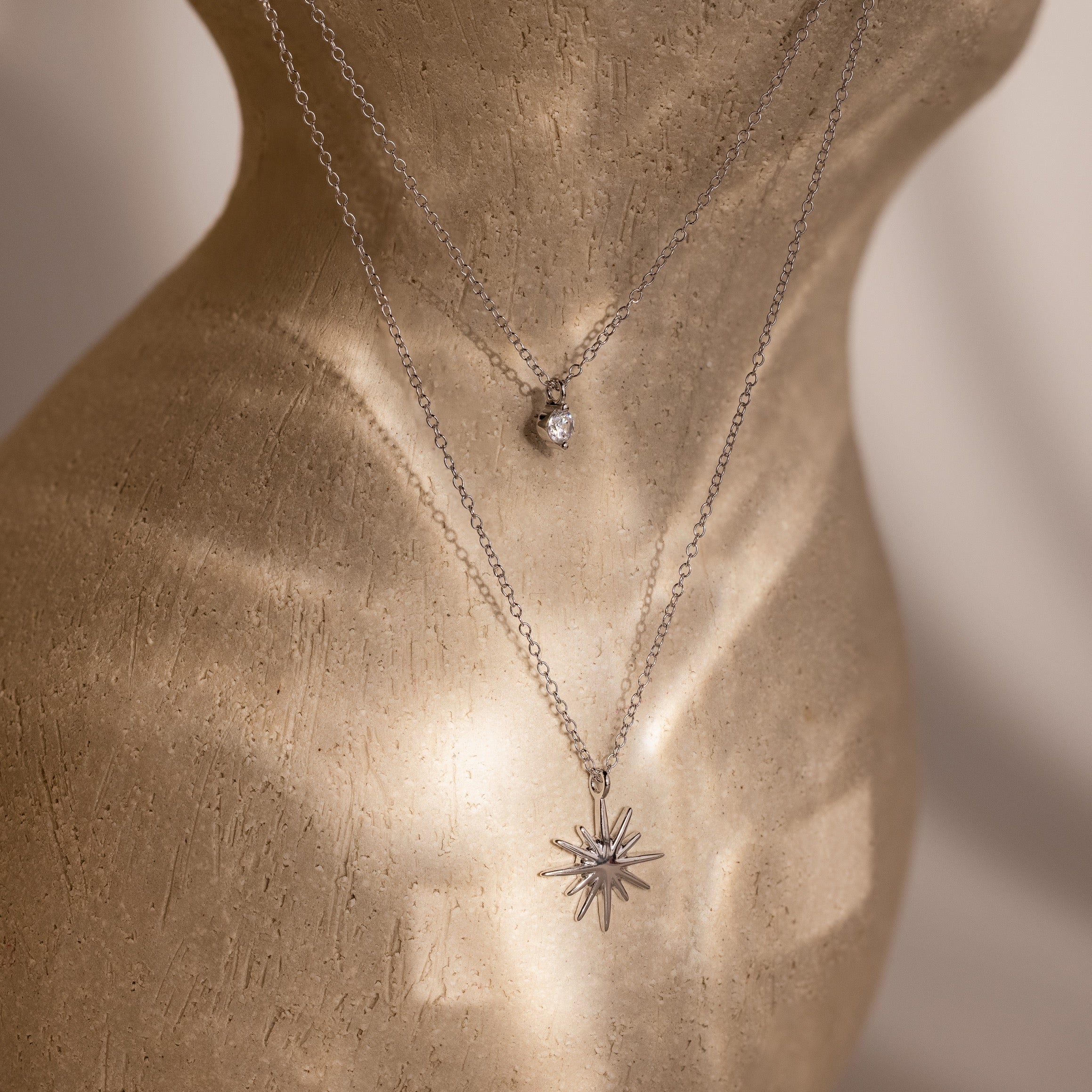 The Laura Starburst Duo Necklace in Silver, shown on a mannequin, features two layered necklaces—one with a diamond accent and the other with a bold starburst pendant.