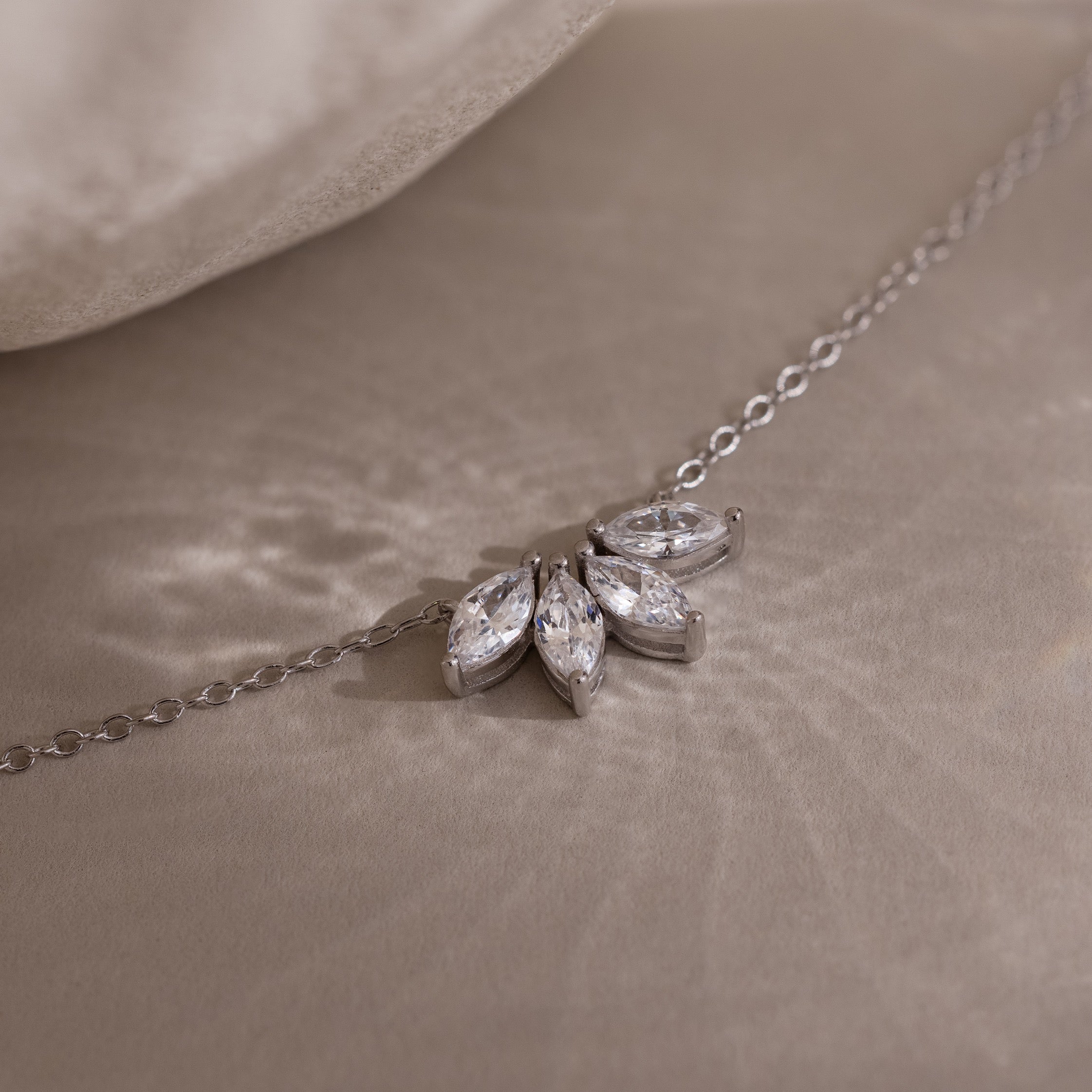 The Marquise Diamond Necklace in Silver, featuring three marquise-cut diamonds arranged in a floral design, rests elegantly on a soft beige surface.
