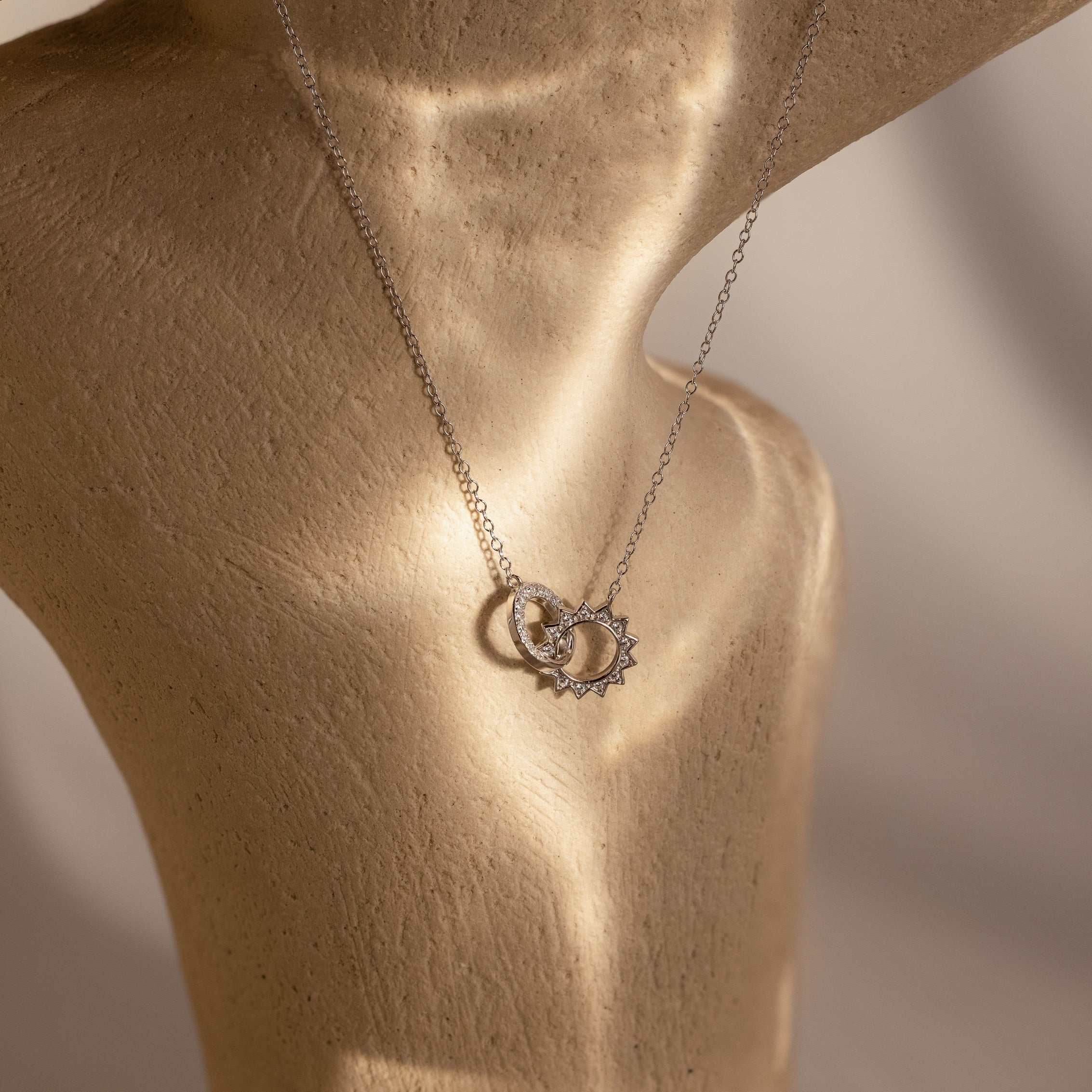 The Sun  Moon Eternity Necklace in Silver features intertwined sun and moon pendants, elegantly displayed on a beige textured mannequin—perfect for any occasion.