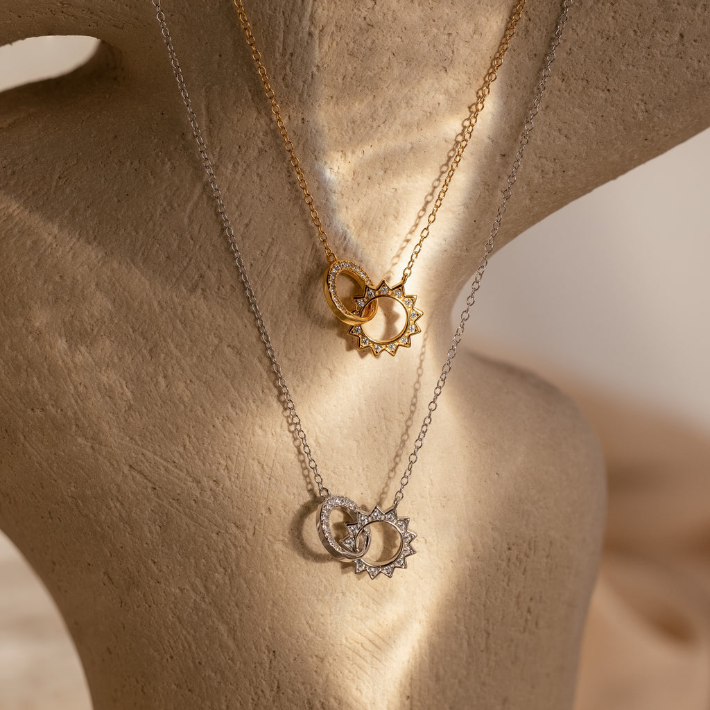 The Sun  Moon Eternity Necklace features gold sun and silver moon pendants, artfully intertwined and elegantly displayed on a textured stand.