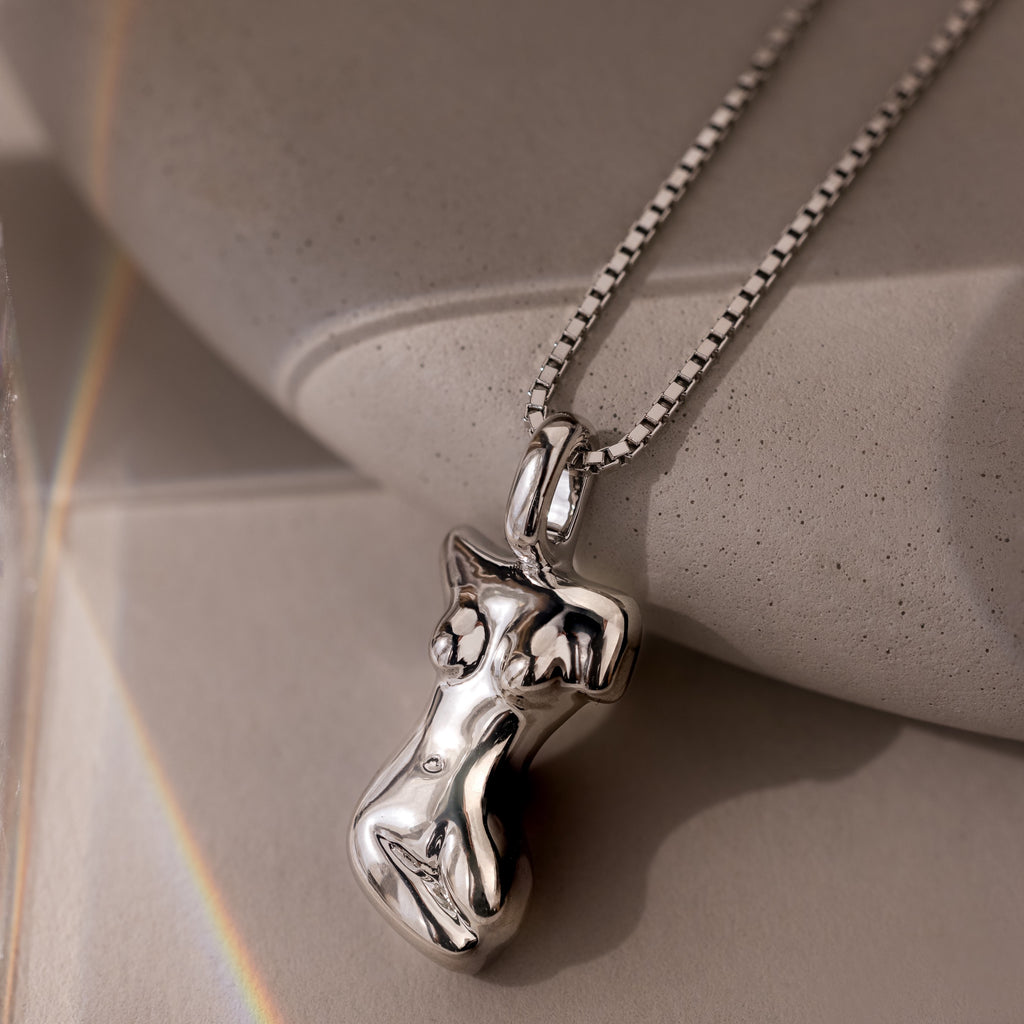 The Kinn Body Necklace in Silver features an abstract female torso pendant and rests on a smooth stone surface—an elegant, empowering jewelry piece.