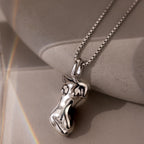 The Kinn Body Necklace in Silver features an abstract female torso pendant and rests on a smooth stone surface—an elegant, empowering jewelry piece.