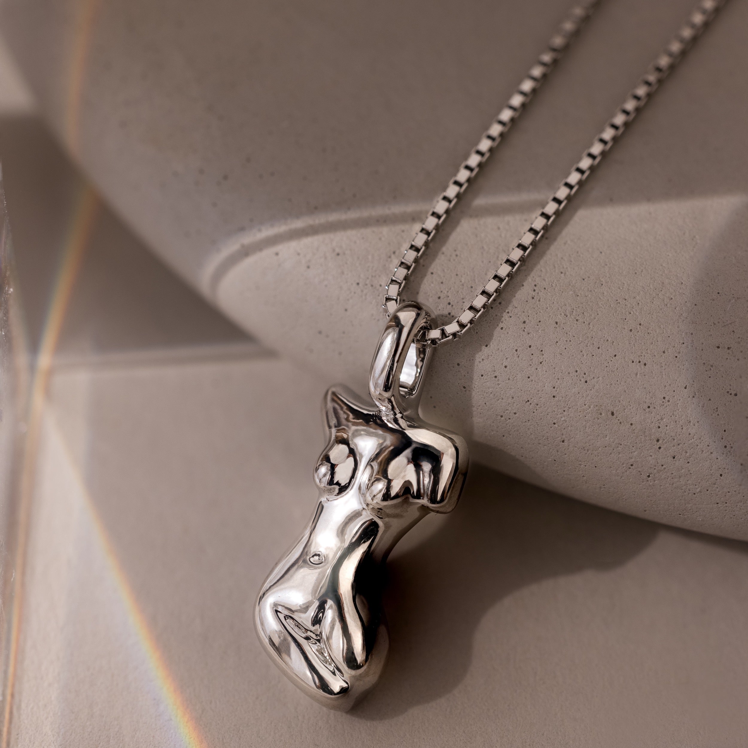 The Kinn Body Necklace in Silver features an abstract female torso pendant and rests on a smooth stone surface—an elegant, empowering jewelry piece.