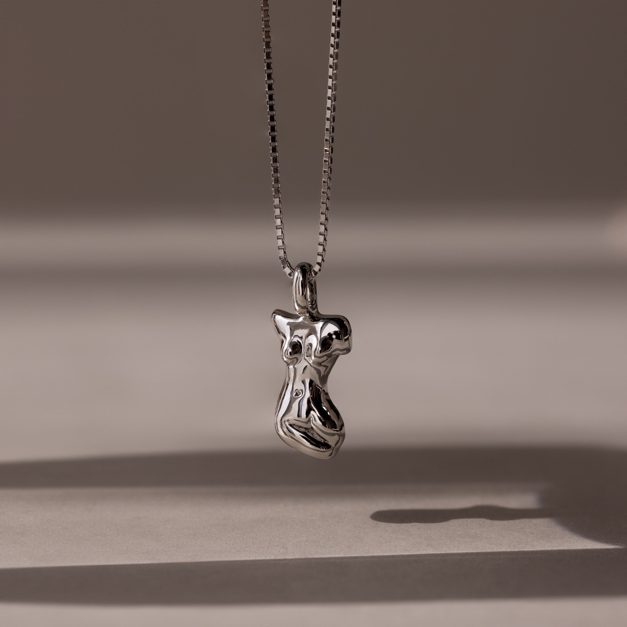 The Kinn Body Necklace in Silver features an abstract pendant on a delicate chain, casting a soft shadow—an elegant and empowering accessory inspired by the Venus de Milo.