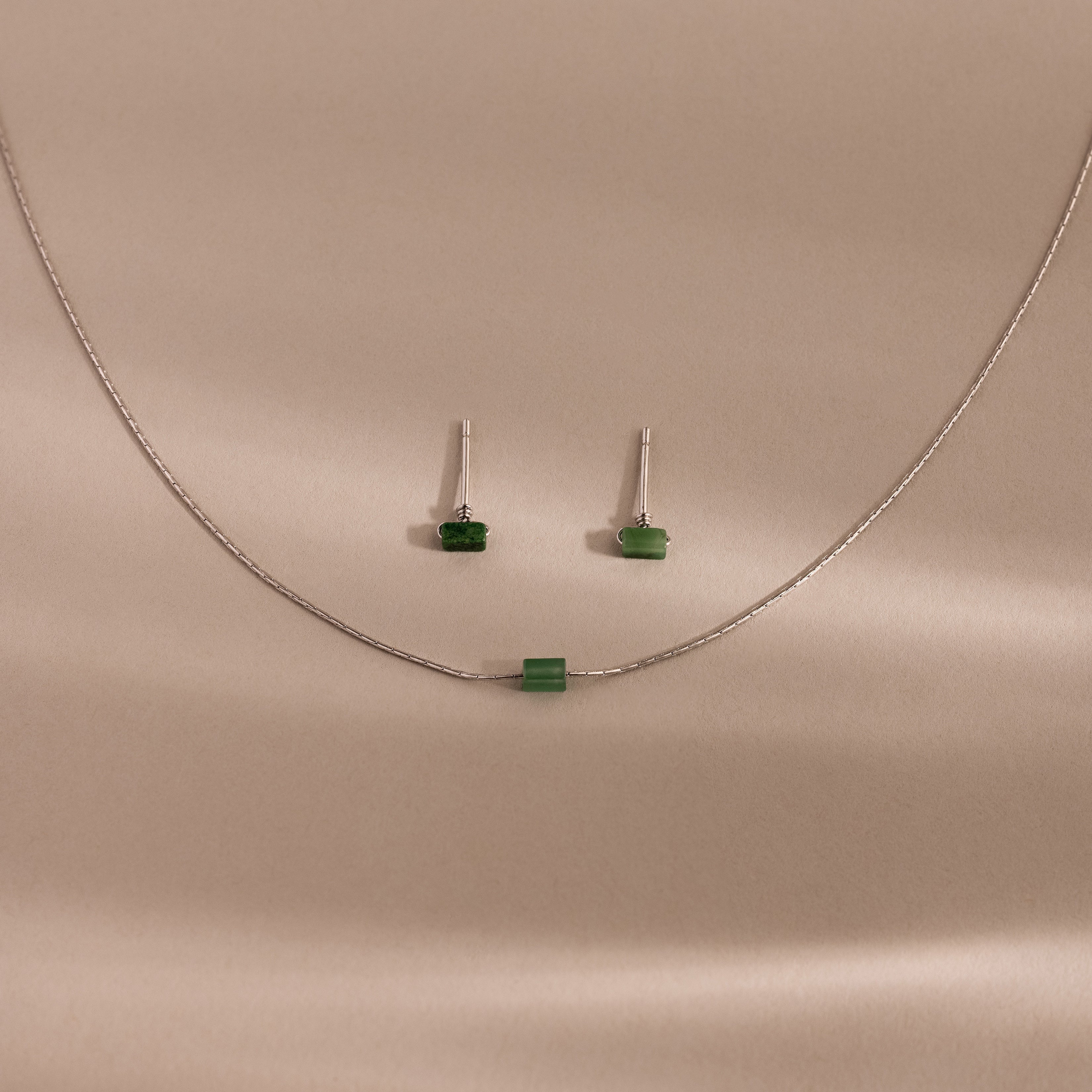 Jade Bar Jewelry Set in Sterling Silver, featuring a silver necklace with a small green bead and matching jade earrings, displayed on a beige background.