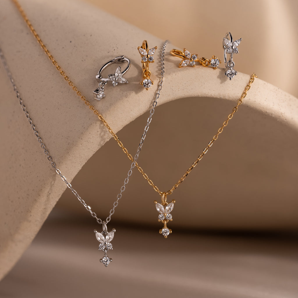The Diamond Butterfly Drop Jewelry Set features gold and silver diamond butterfly designs with necklaces, huggie earrings, and rings displayed on a beige surface.
