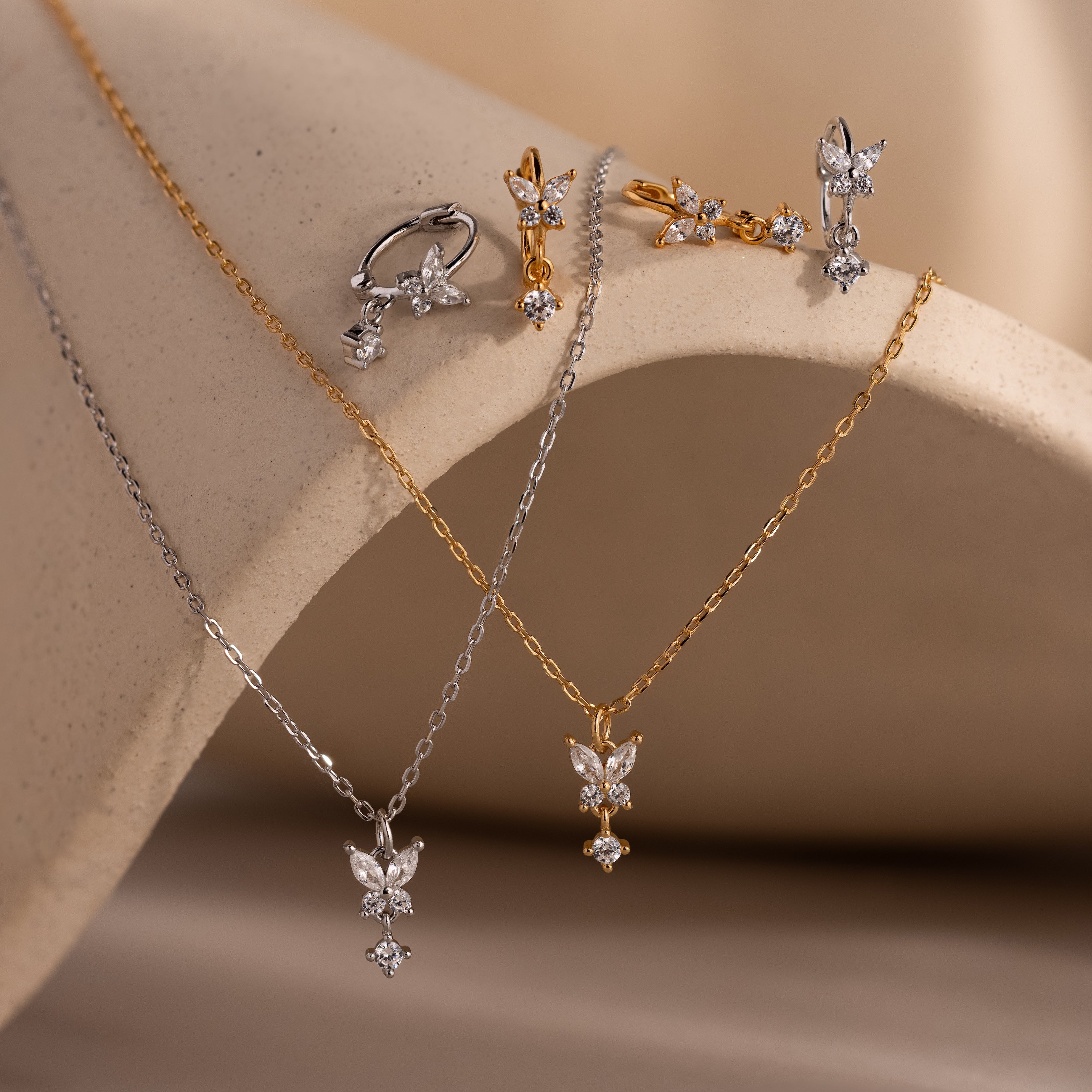 The Diamond Butterfly Drop Jewelry Set features gold and silver diamond butterfly designs with necklaces, huggie earrings, and rings displayed on a beige surface.
