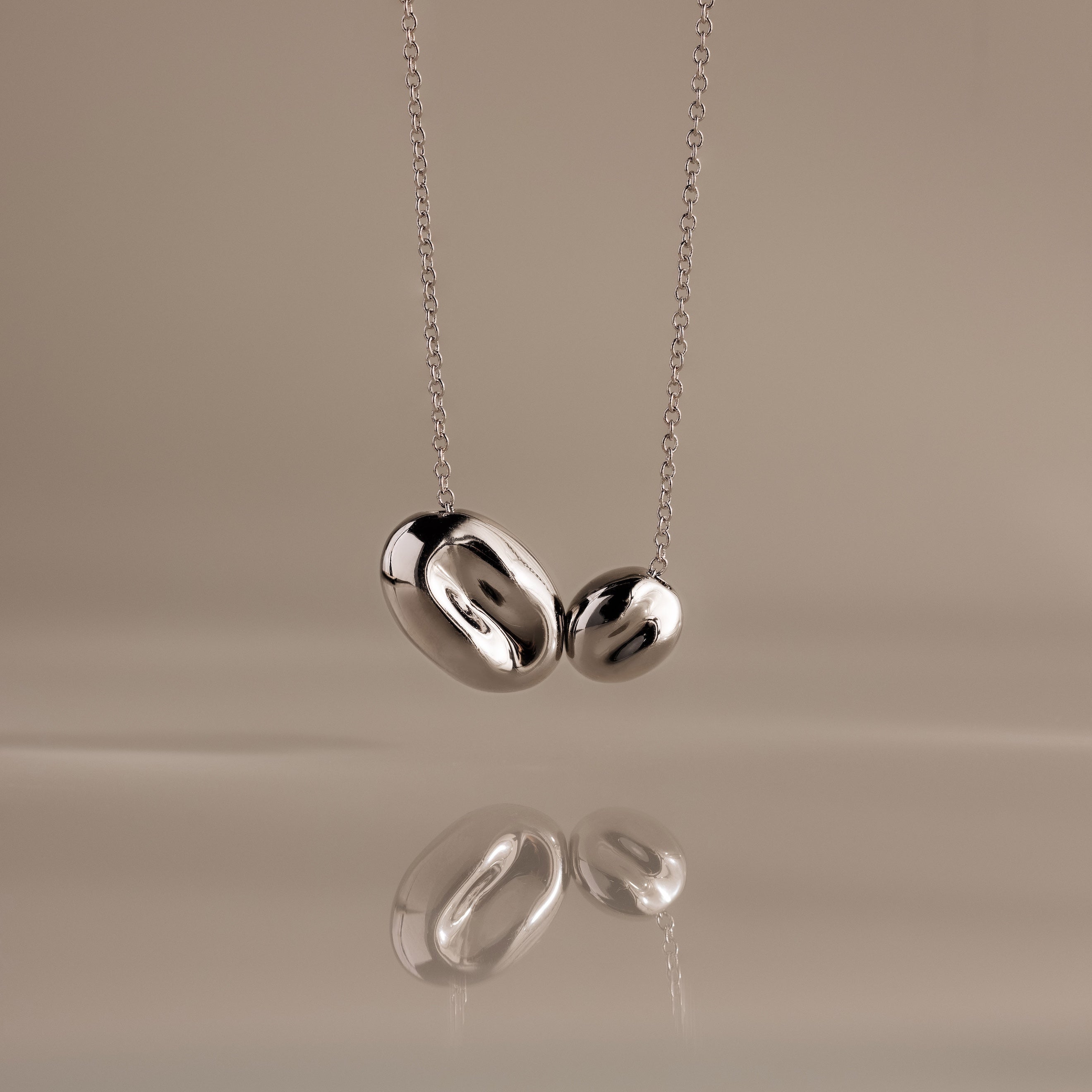 The Duo Pebble Necklace in Silver features two smooth, oval pendants, elegantly displayed against a beige background with a subtle reflection beneath.