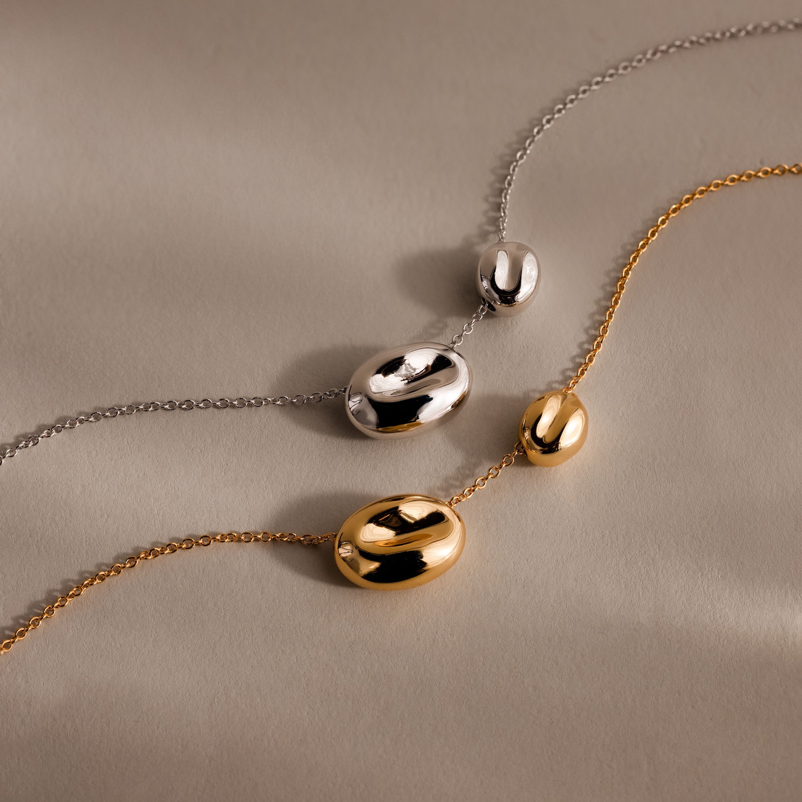 The Duo Pebble Necklace features two chain necklaces, one silver and one gold, each adorned with a smooth pebble pendant, displayed elegantly on a beige surface.