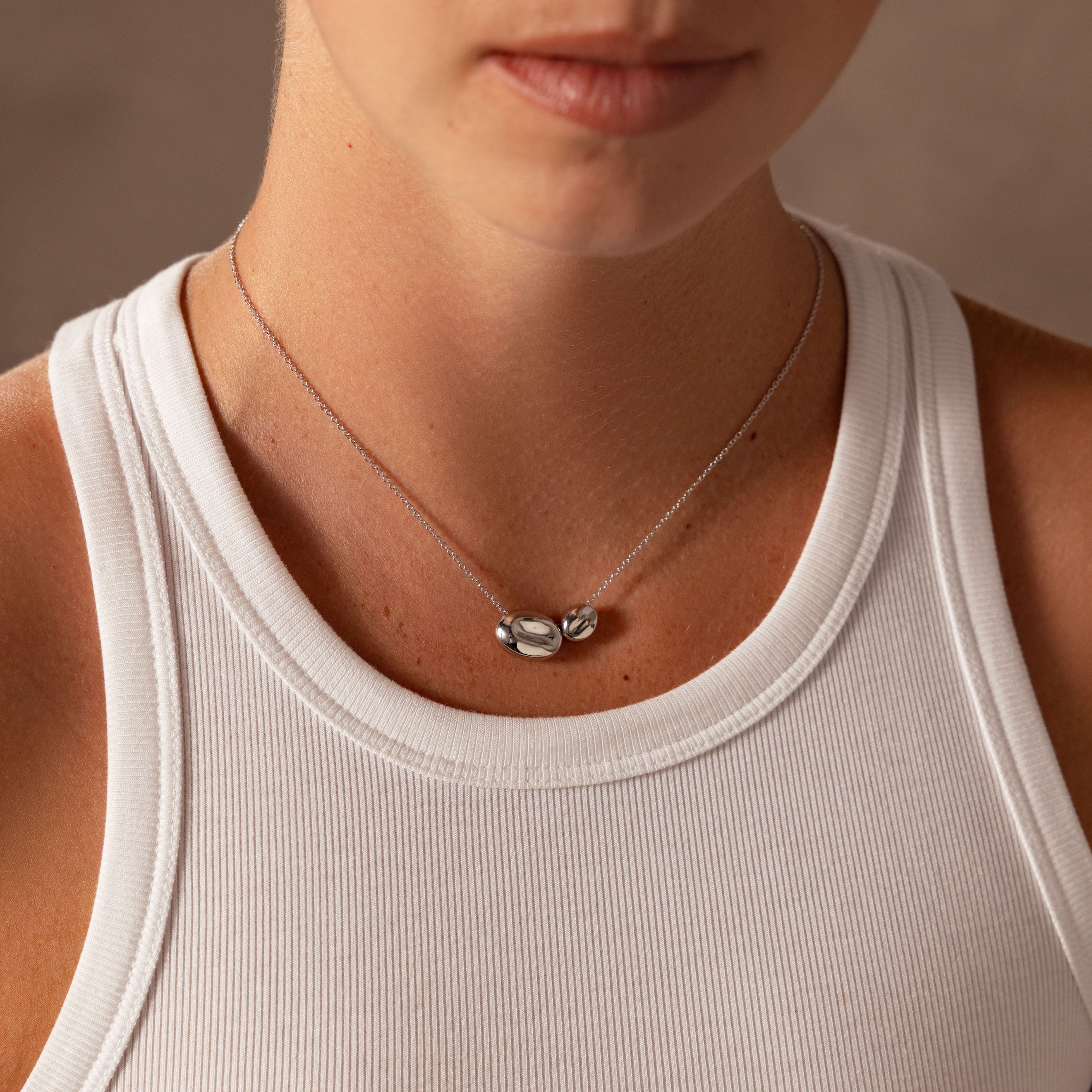A person in a white tank top wears the Duo Pebble Necklace in Silver, featuring two oval pendants. Please provide a product description to help identify SEO keywords.