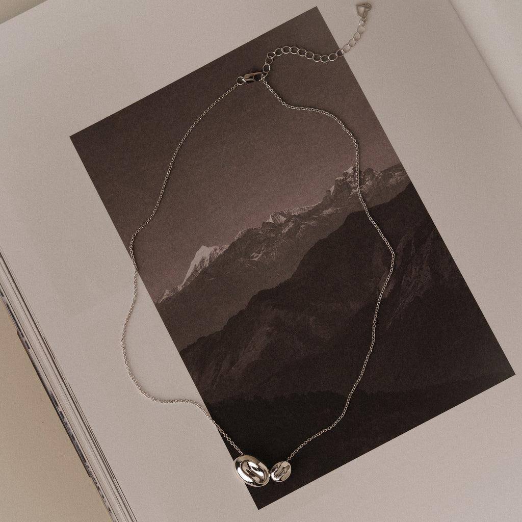 The Duo Pebble Necklace in Silver, featuring two dainty pendants, rests on an open book with a black and white mountain photo, lending subtle elegance to the scene.